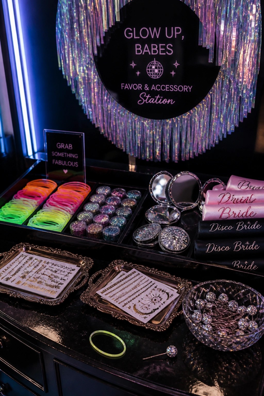 A black table holds stacks of neon glow bracelets, round glitter compacts, pink satin sashes, gold temporary tattoos on trays, and a bowl of jeweled hair pins beneath a circular disco sign.