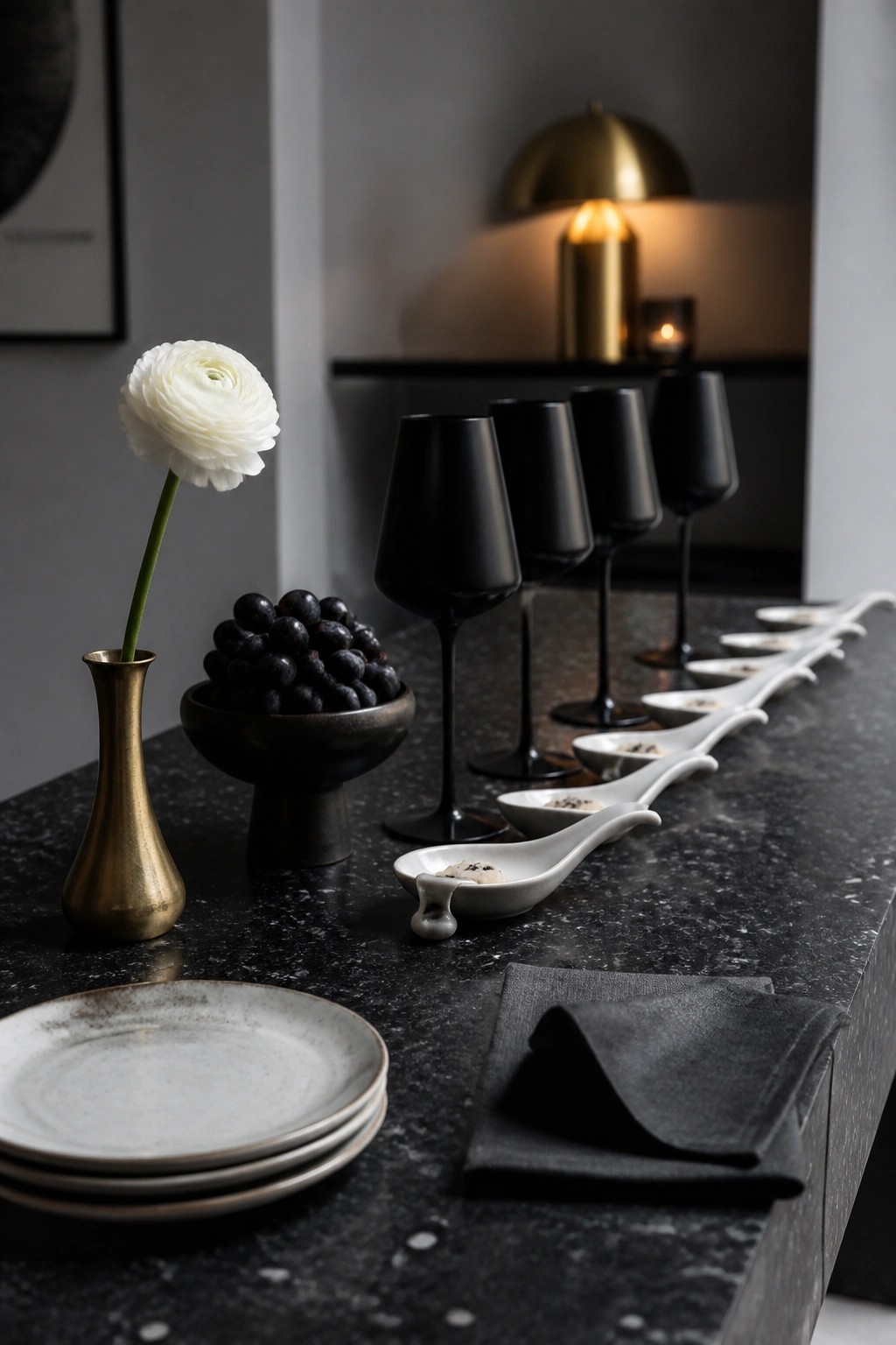 A dark marble table holds a row of matte black wine glasses beside white ceramic tasting spoons, with a brass vase and bowl of dark grapes nearby.