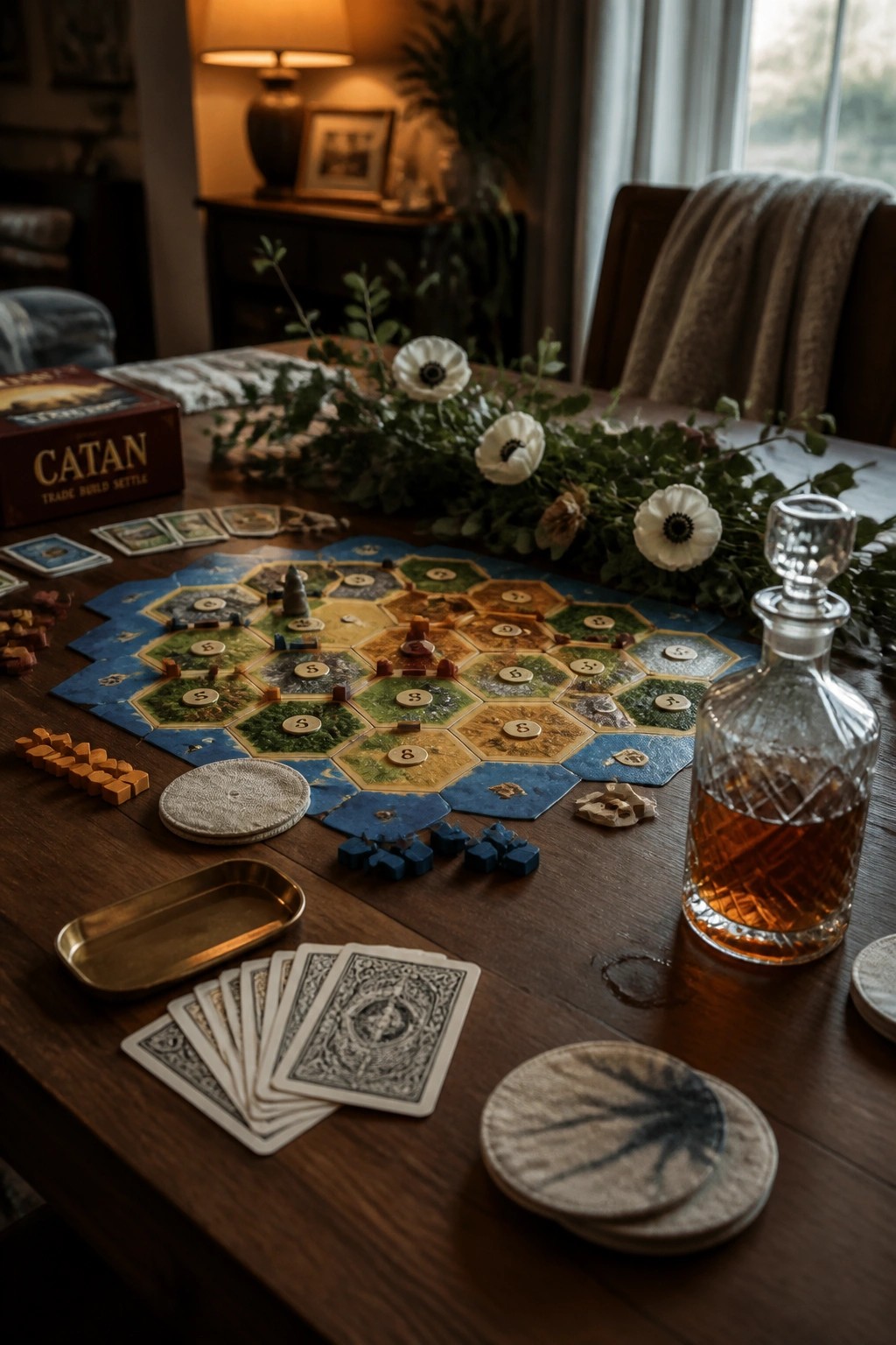 A wooden table holds a fully laid out Catan board game with player pieces, resource cards, a glass decanter of amber liquid, and a few floral stems scattered across the surface.